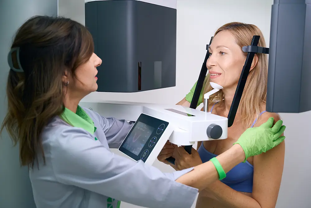 An orthodontist positions a patient in a CBCT scanning machine, adjusting the equipment while the patient stands with their head secured for imaging.