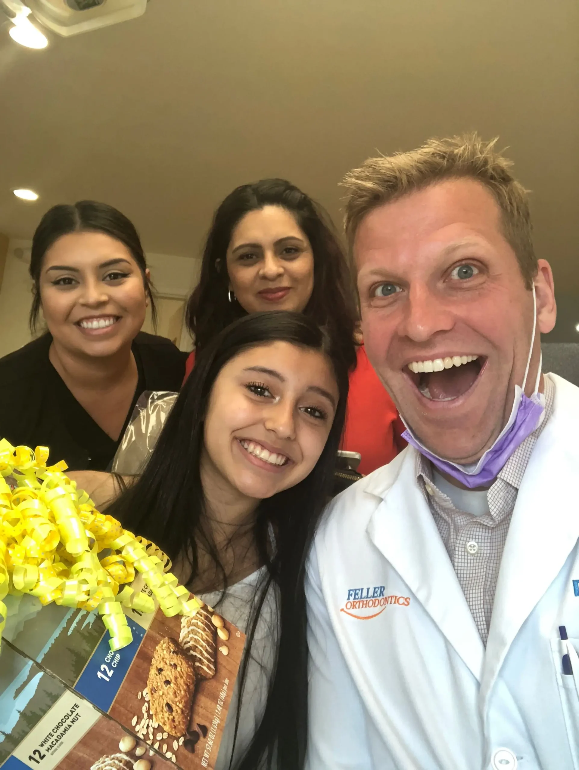 Dr. Feller and nurse smiling together with a patient and parents indoors as one person holds a gift box with cookies, while another wears a white coat labeled “Feller Orthodontics” with a purple mask resting below the chin.