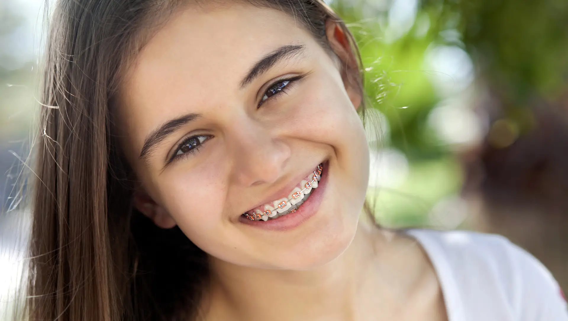 Smiling Teen Girl with Colorful Braces A young teenage girl with long brown hair smiles outdoors, showing colorful orthodontic braces in bright natural sunlight.