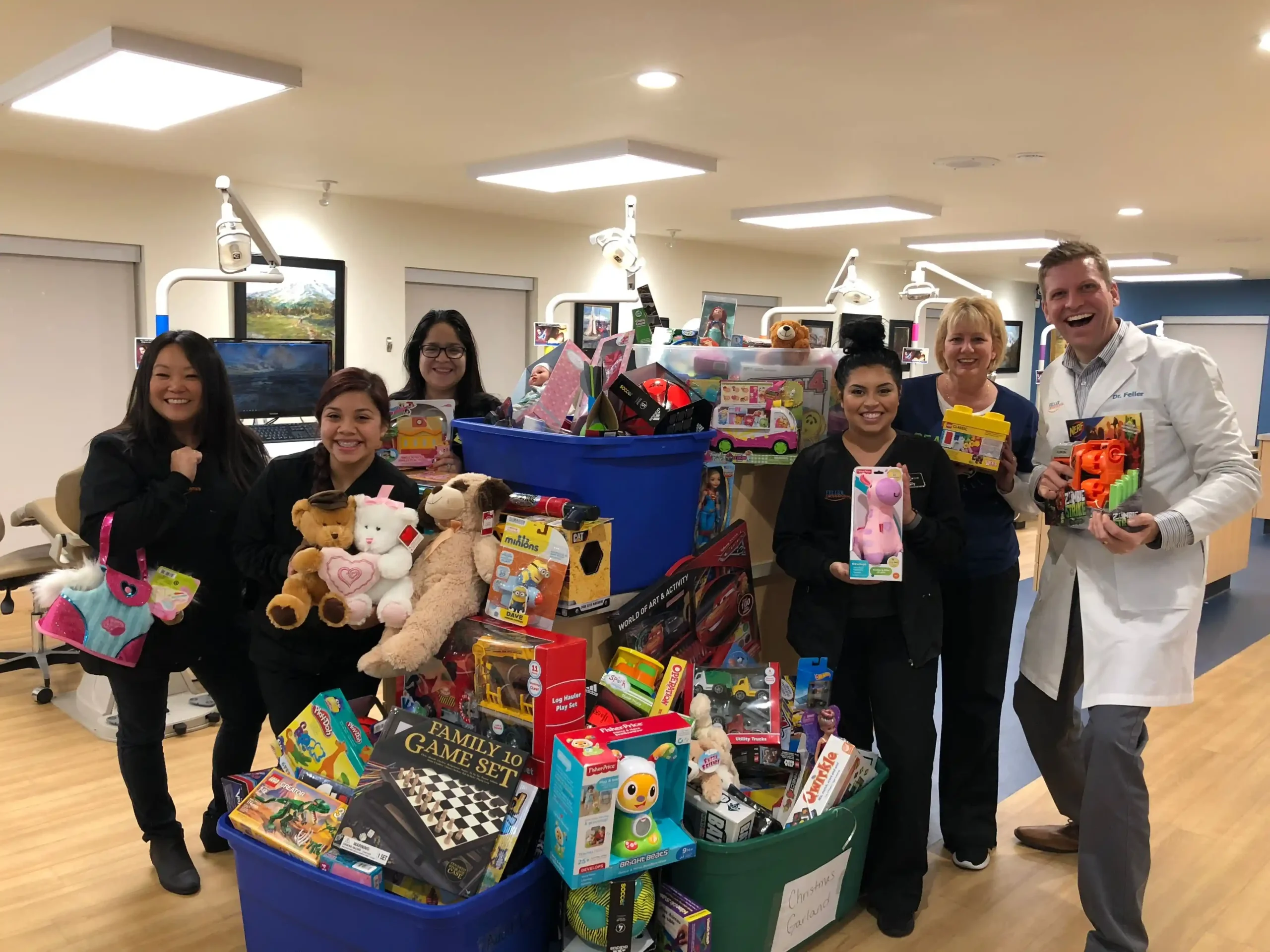 Dr. Feller and Team Collect Toys for Holiday Giveaway Dr. Feller and team members stand together indoors, smiling as they hold toys and gather around large bins filled with donated items for a holiday giveaway.
