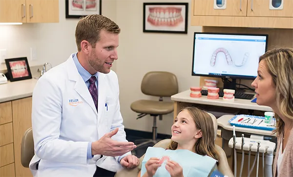 Orthodontist Dr. Feller speaks with a smiling child seated in an exam chair while a mother looks on, with tools, models, and a monitor displaying teeth visible in the background.